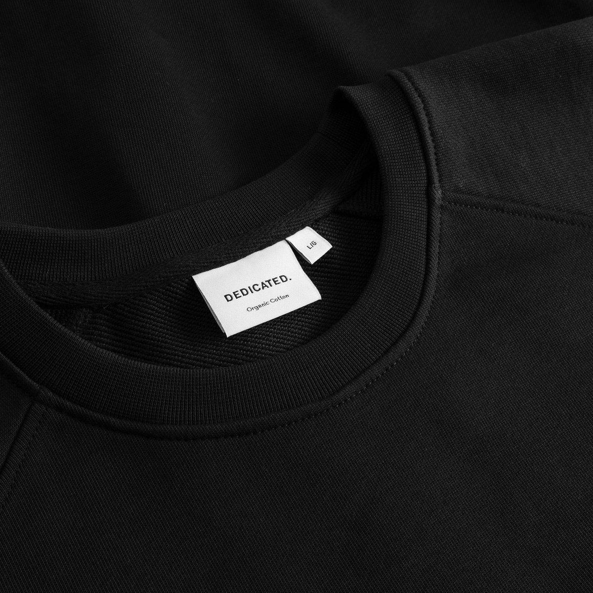 A close-up of the DEDICATED. brand label and ribbed neckline of the black Sweatshirt Malmoe Perch EMB.