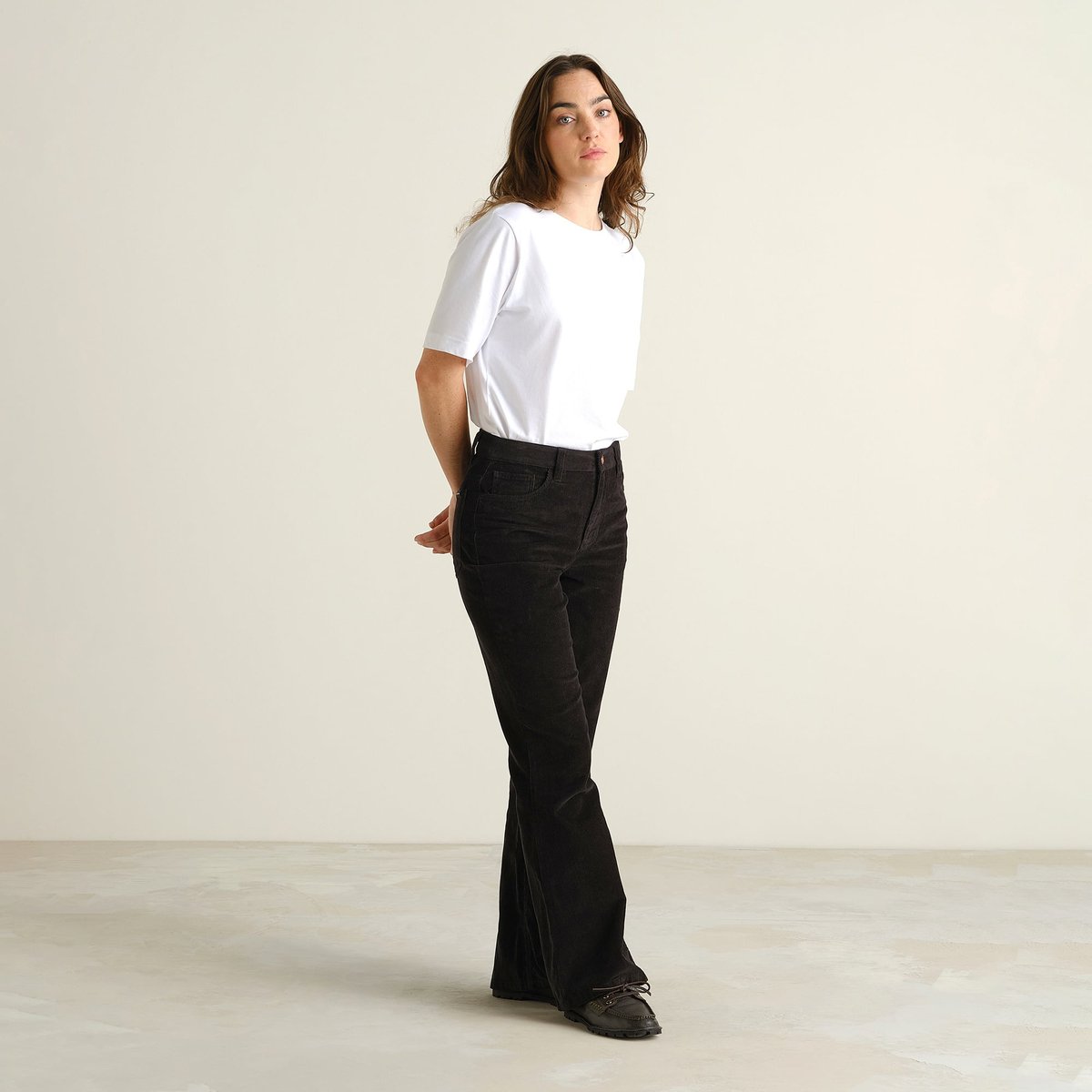 Model in der schwarzen Bootcut-Cordhose Ryda, kombiniert mit einem weißen T-Shirt und schwarzen Loafern.