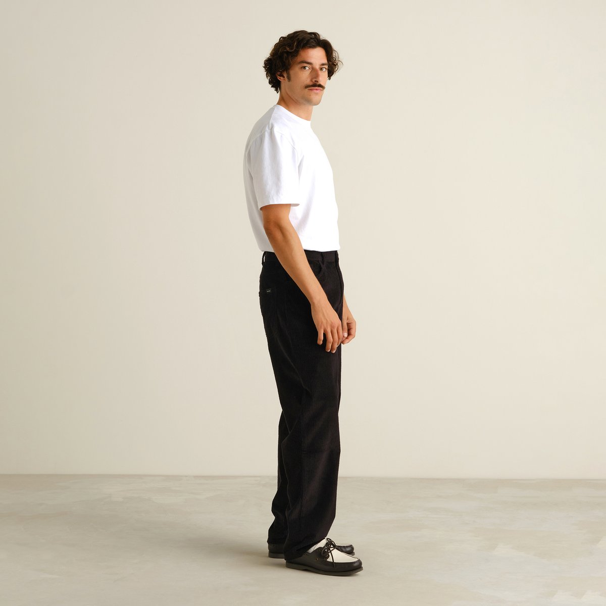 A model in a studio shows off the relaxed fit of the black Hamburg Corduroy Pants, styled with a classic white tee.