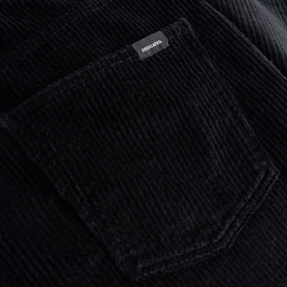 A close-up shot of the rich black corduroy fabric and back pocket of the Hamburg pants, showing the Dedicated brand label.