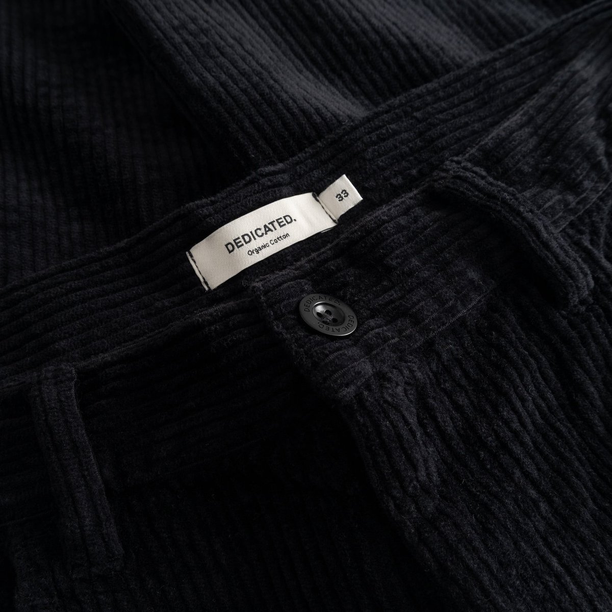 Close-up of the waistband on the black Hamburg Corduroy Pants, showing the Dedicated brand label and button detail.