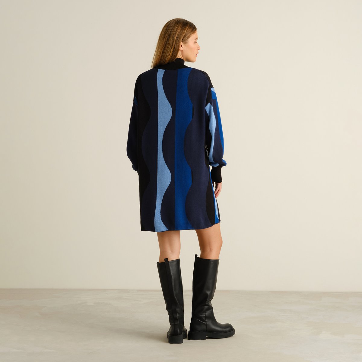Back view of a model wearing the Nikkala dress in a blue wavy stripe pattern, styled with black knee-high boots.