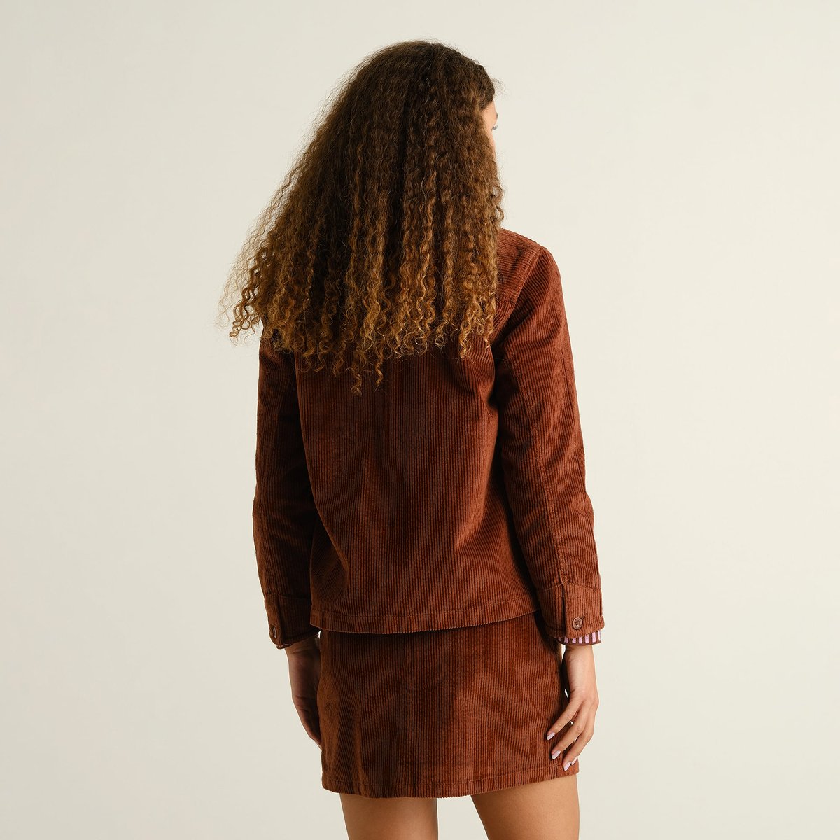 Back view of the Stiby Corduroy Jacket in Cappuccino Brown, worn as a set with the matching skirt by a model with curly hair.