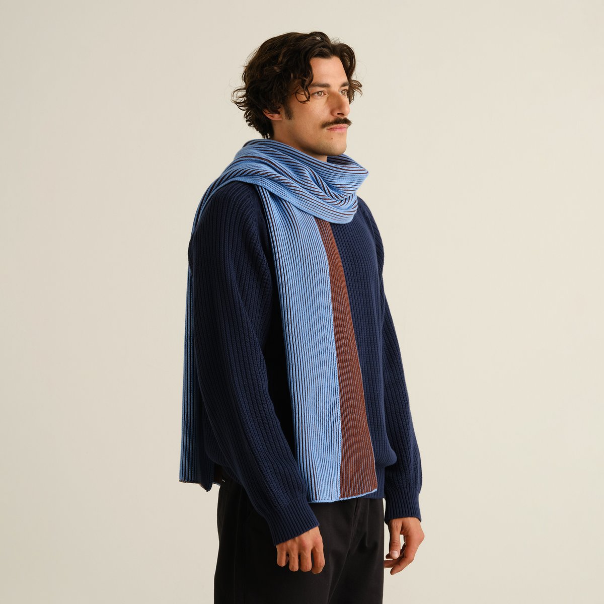 Man i profil bär den randiga halsduken Norrfors i Cappuccino Brown och Cornflower Blue till en marinblå stickad tröja.