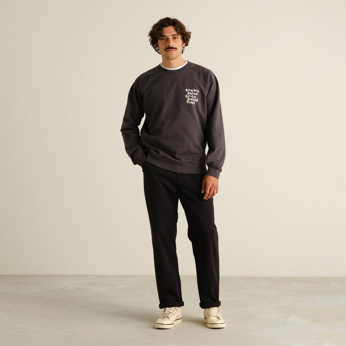 A man with a mustache wearing black Hudiksvall chinos, a dark grey sweatshirt, and off-white sneakers against a background.
