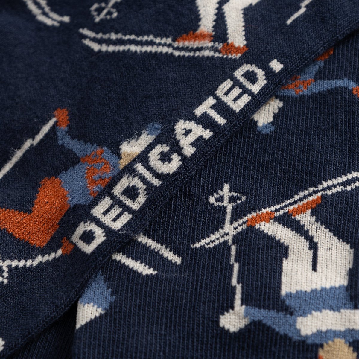 A close-up of the Dedicated Sigtuna socks in Estate Blue, showing the detailed knitted pattern of skiers and the brand logo.