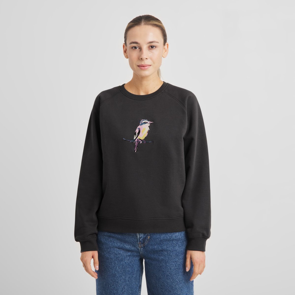 A model seen from the front wears the Ystad sweatshirt in charcoal with a colourful painted bird embroidery.