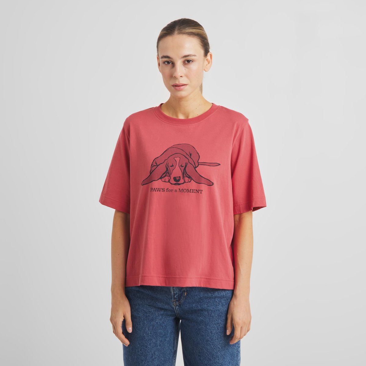 Woman wearing a holly red t-shirt with a print of a sleeping dog and the text 'Paws for a moment', styled with blue jeans.