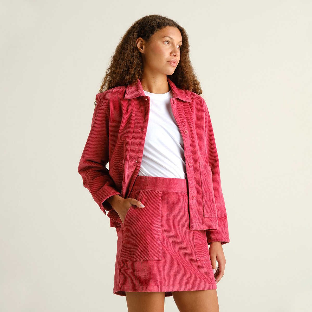 A woman with curly hair models a matching set of a Holly Red corduroy jacket and the Majorna skirt, over a white t-shirt.