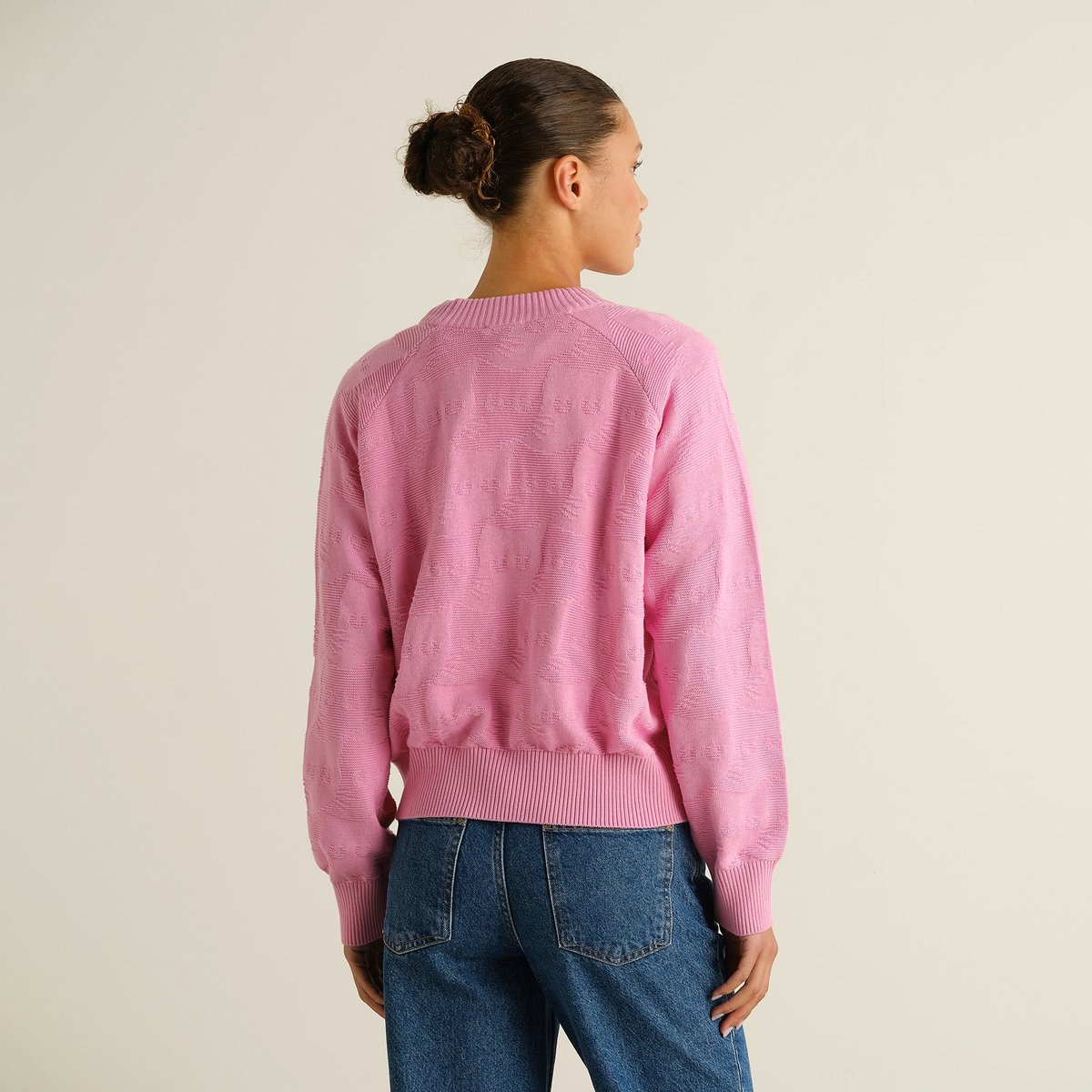Woman seen from the back wearing the pink Husie Pepita Cats Knit sweater paired with blue jeans in a studio.