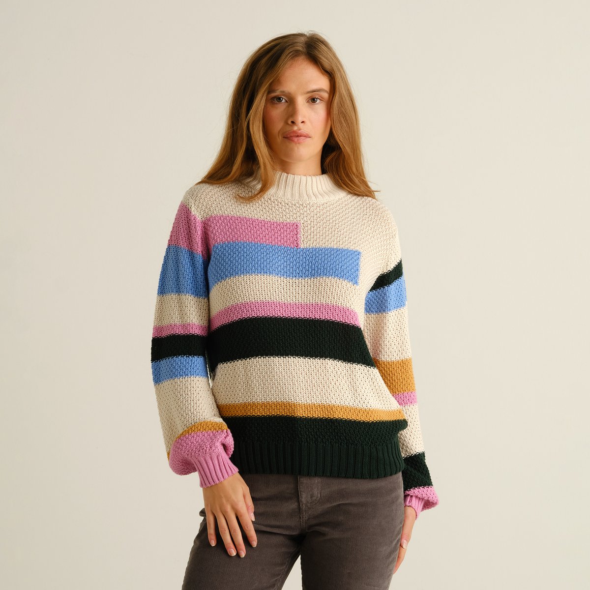 A model wears the chunky Gotland sweater, featuring playful, multi-colored offset stripes, against a simple studio backdrop.