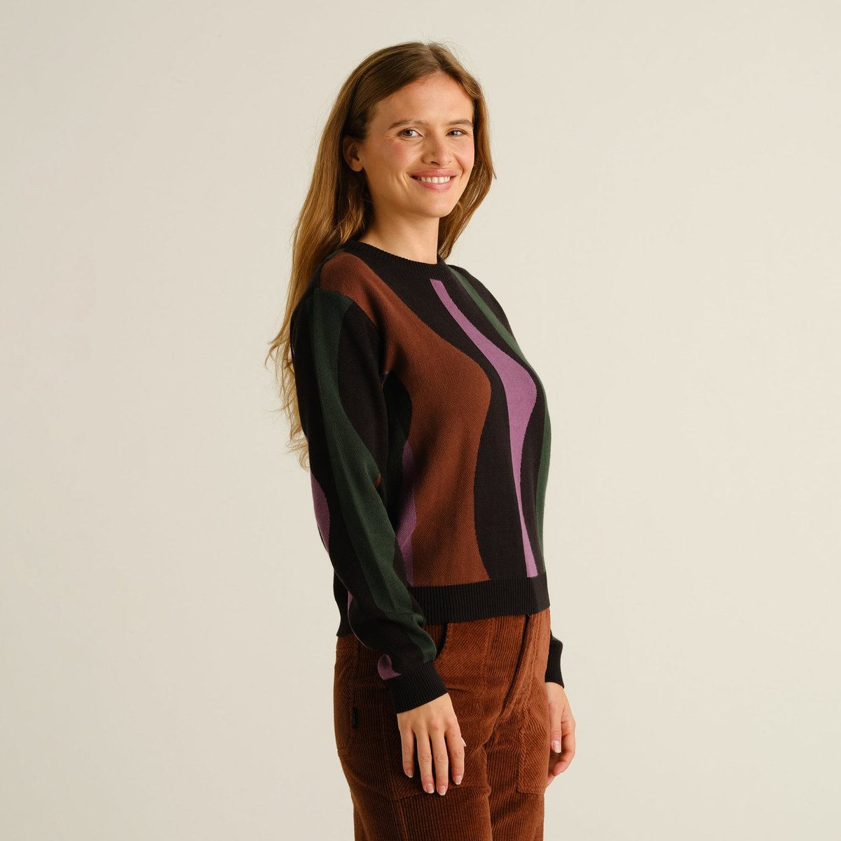 Side view of a smiling woman wearing the Sweater Arendal Wavy Stripe in Multi Color with brown corduroy trousers.