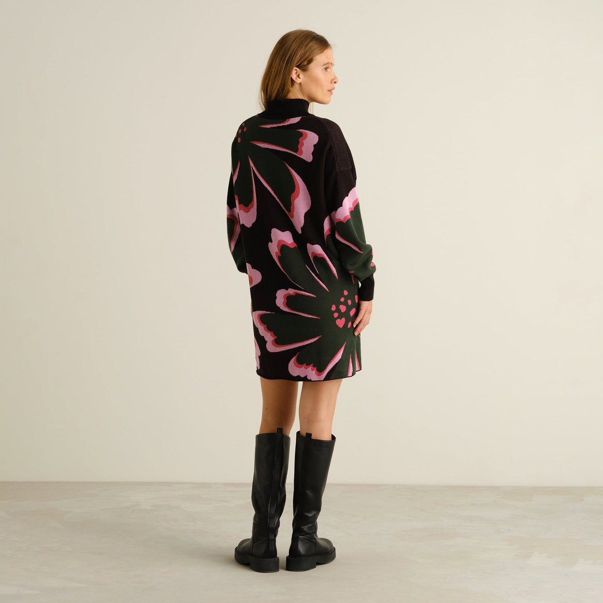Das Strickkleid Nikkala Big Bloom von hinten, getragen von einem Model mit schicken schwarzen, kniehohen Stiefeln.