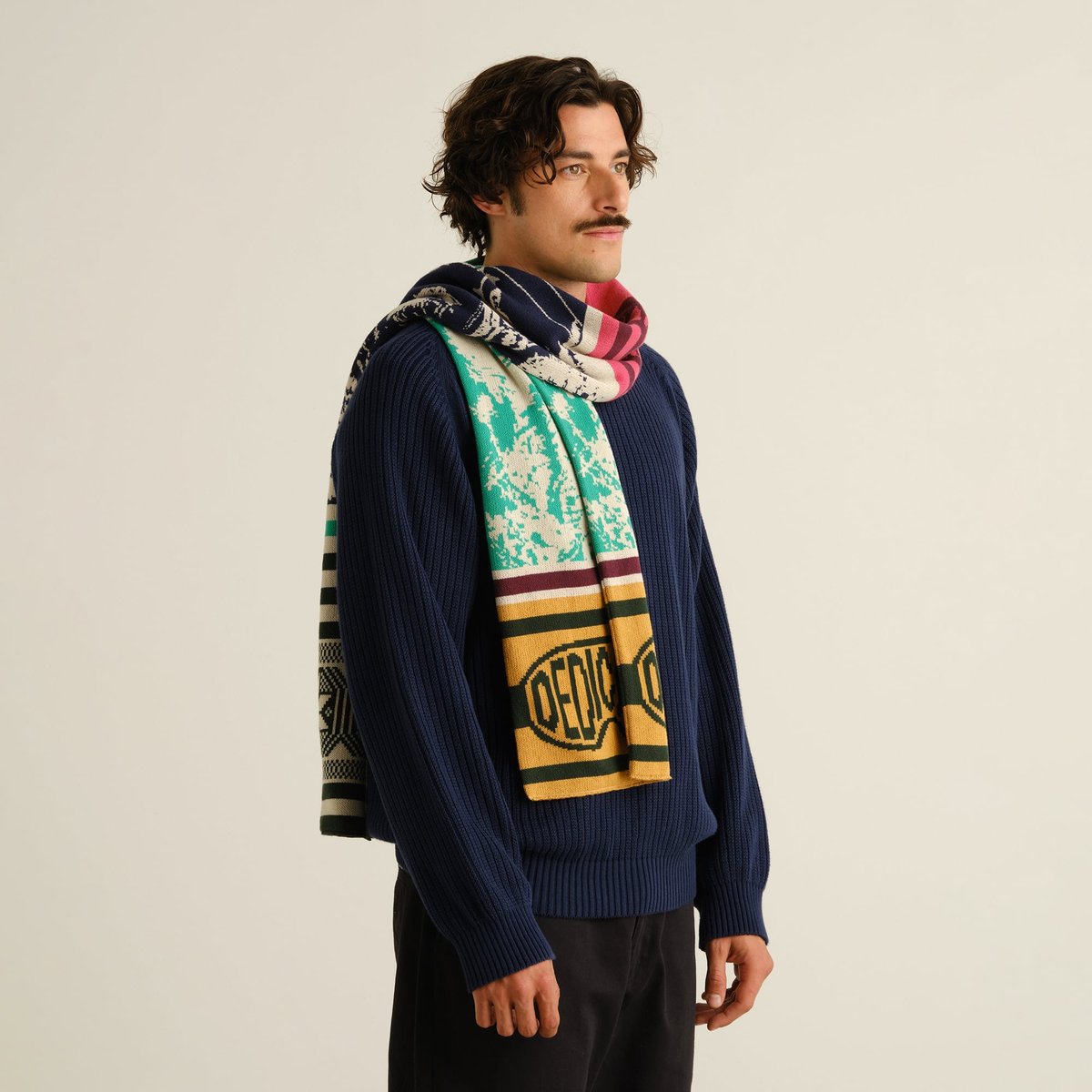 Male model in a studio setting, wearing a navy blue sweater and the vibrant, multi-colored Rossberga Retro Ski scarf.