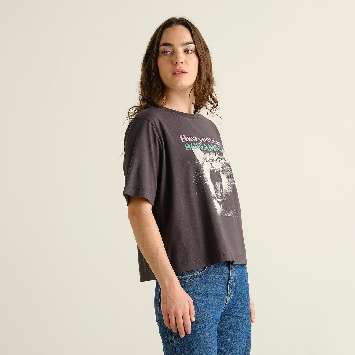 A woman looks over her shoulder, wearing the charcoal T-shirt Vadstena with a Screaming Cat print and a pair of blue jeans.