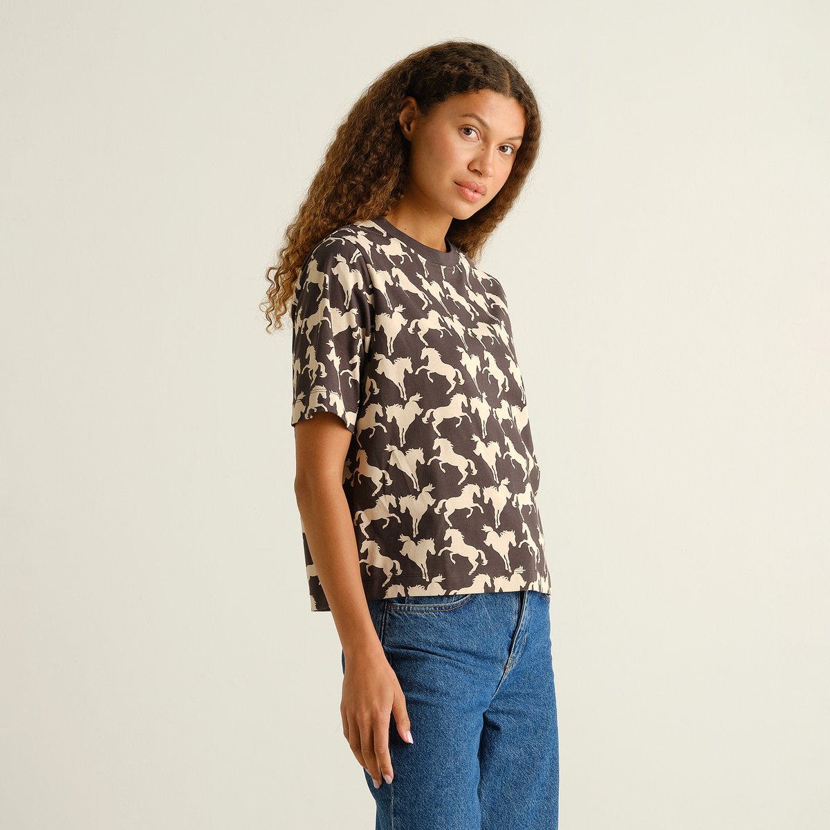 A woman wears the charcoal T-shirt Vadstena with an all-over wild horses print, paired with classic blue jeans.