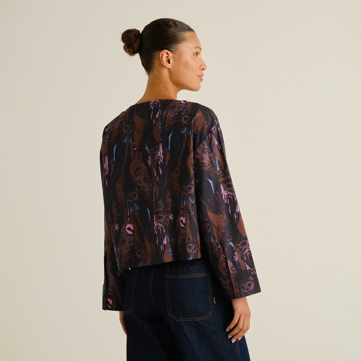 A woman with her hair in a bun shows the back of the Top Udda, a boxy blouse with a vibrant owl print.