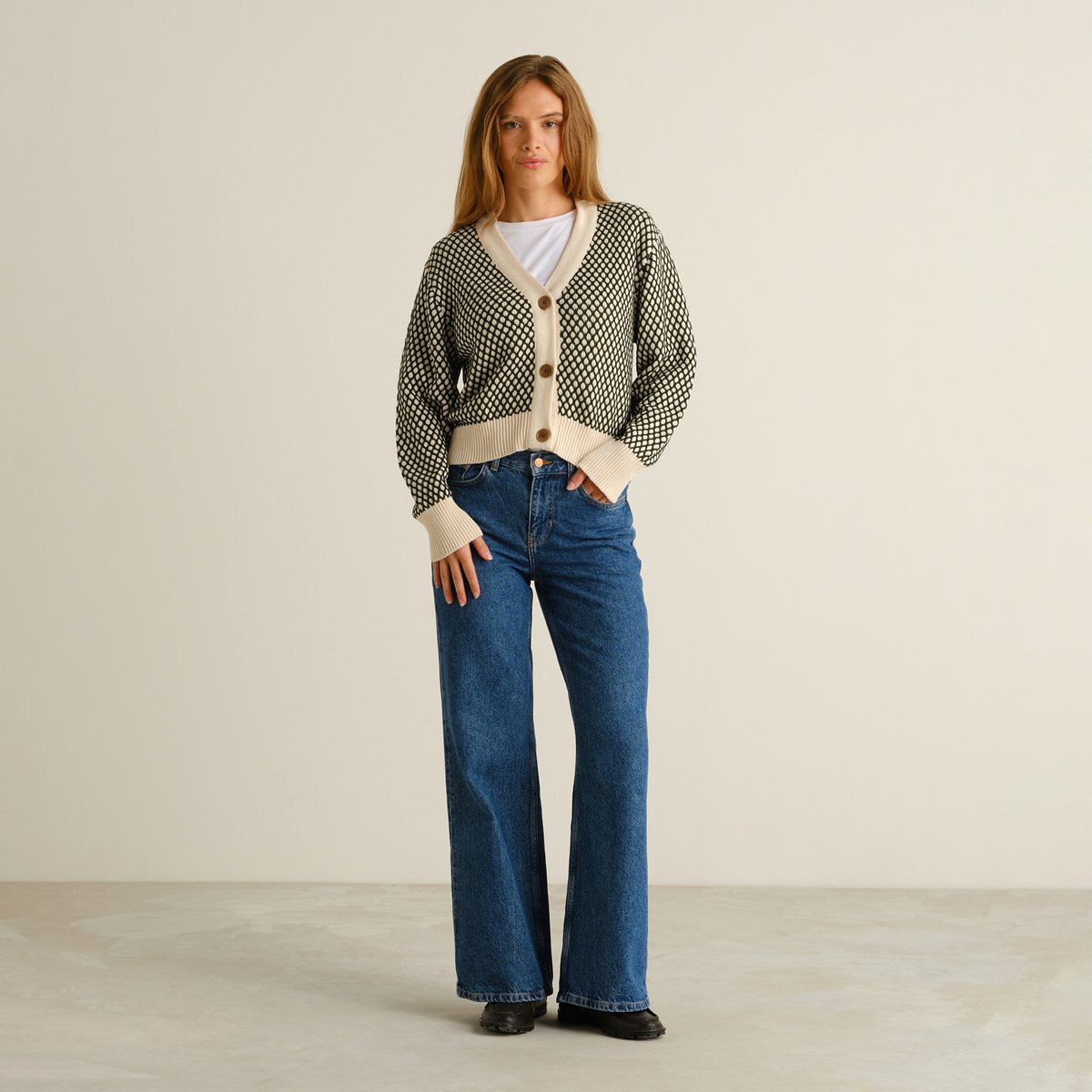Helkroppsbild av en kvinna som bär den mörkgröna och vaniljfärgade stickade koftan Kanan Bubble Knit med jeans med vida ben.