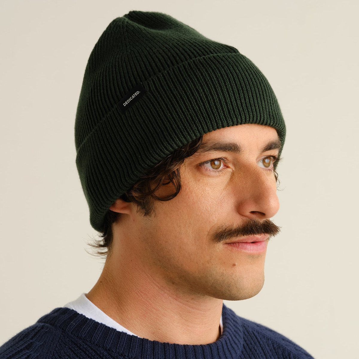 A model looks off-camera, showing off the cozy fit of the dark green ribbed Beanie Hemavan.