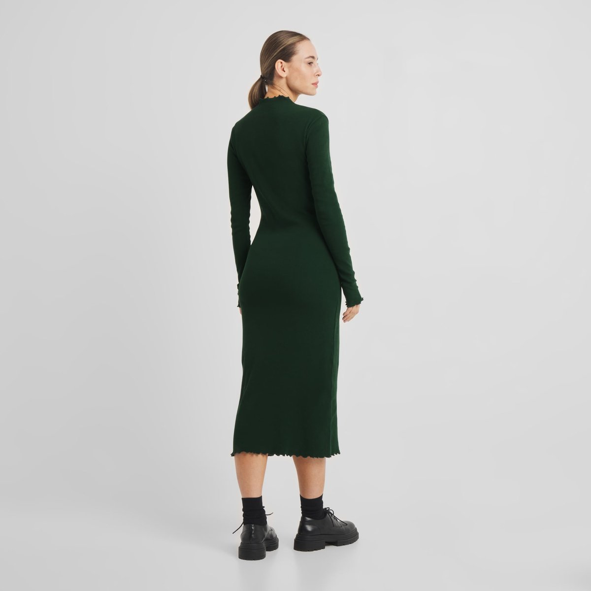 Model from behind wearing the long-sleeved Rib Dress Lofta in dark green, paired with black socks and chunky shoes.