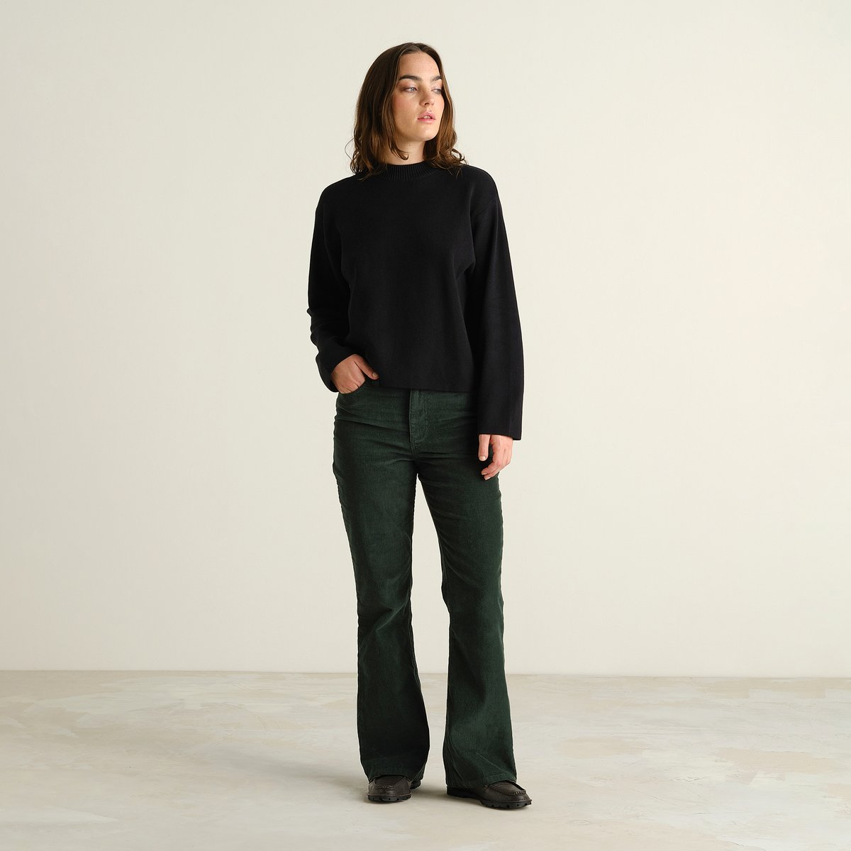 A model wears the dark green Ryda Corduroy Pants, a modern bootcut style, paired with a classic black knit sweater.