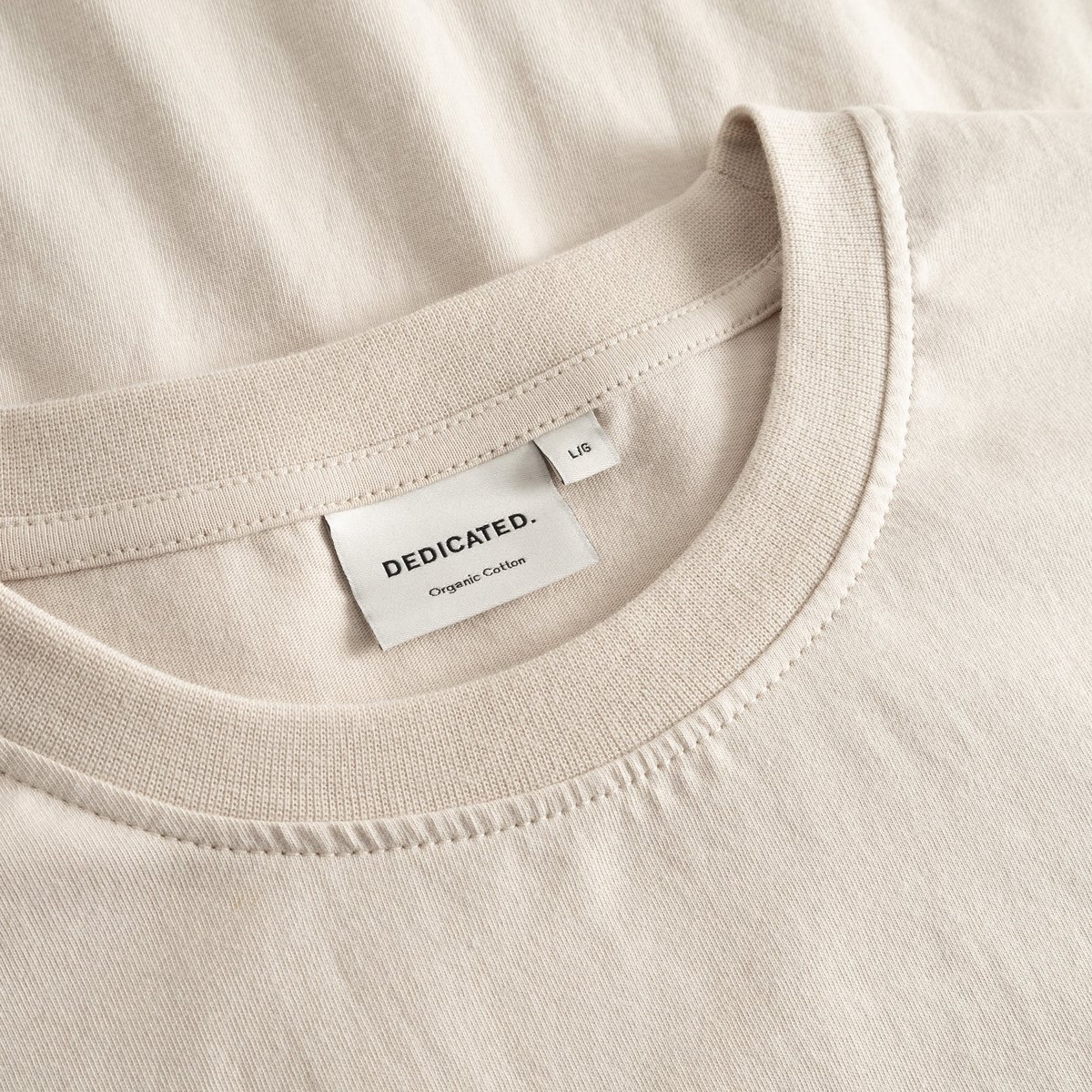 Close-up of the Dedicated brand label and ribbed collar on the classic Stockholm t-shirt in a soft oat white.
