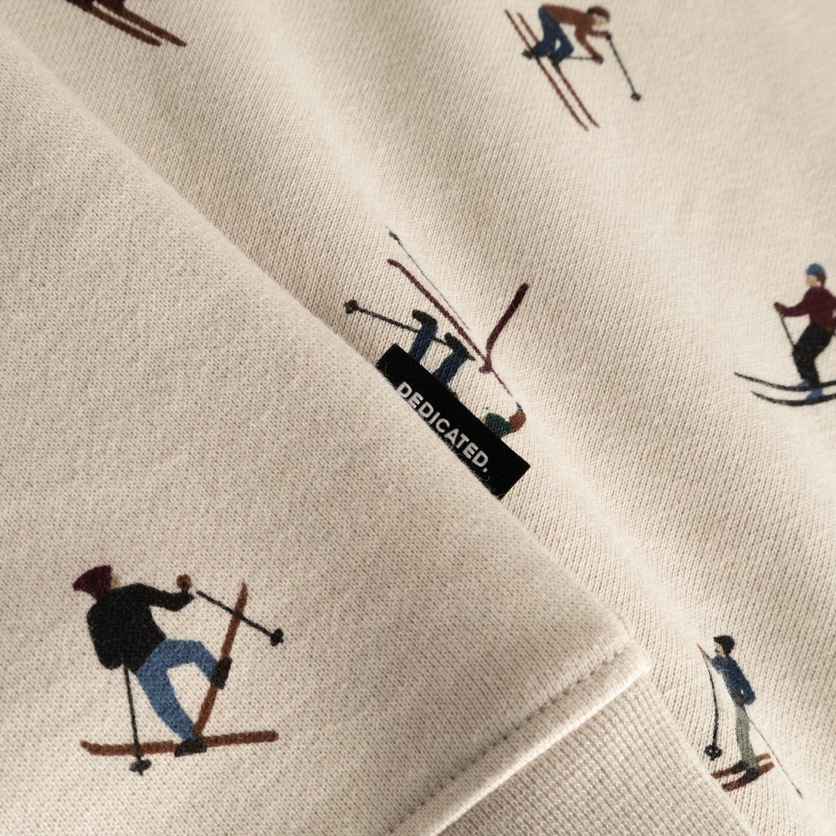 Close-up detail shot of the oat white Malmoe Ski Team sweatshirt fabric, showing the fun skier print and Dedicated brand tab.