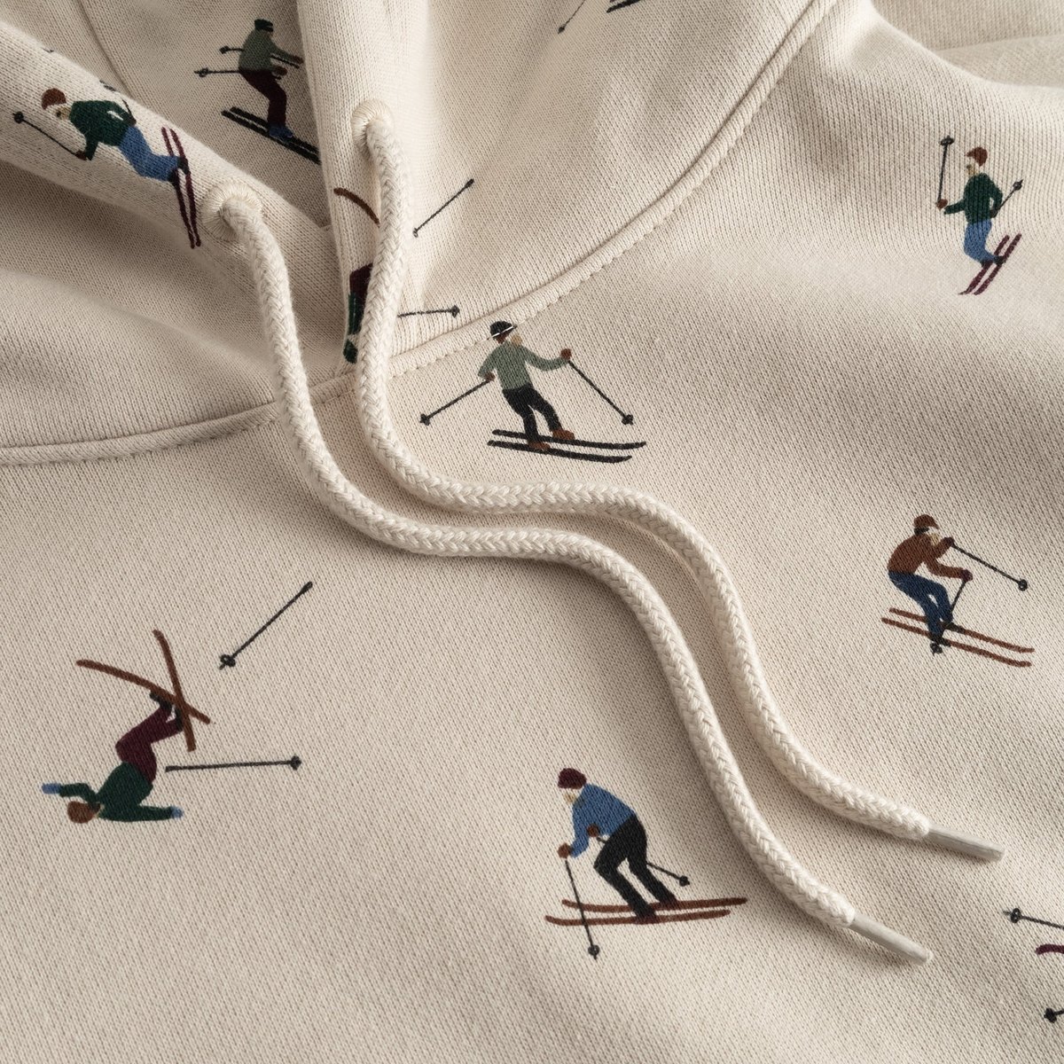 Close-up detail of the oat white Falun Ski Team hoodie, showing the playful all-over embroidered skier print and drawstrings.
