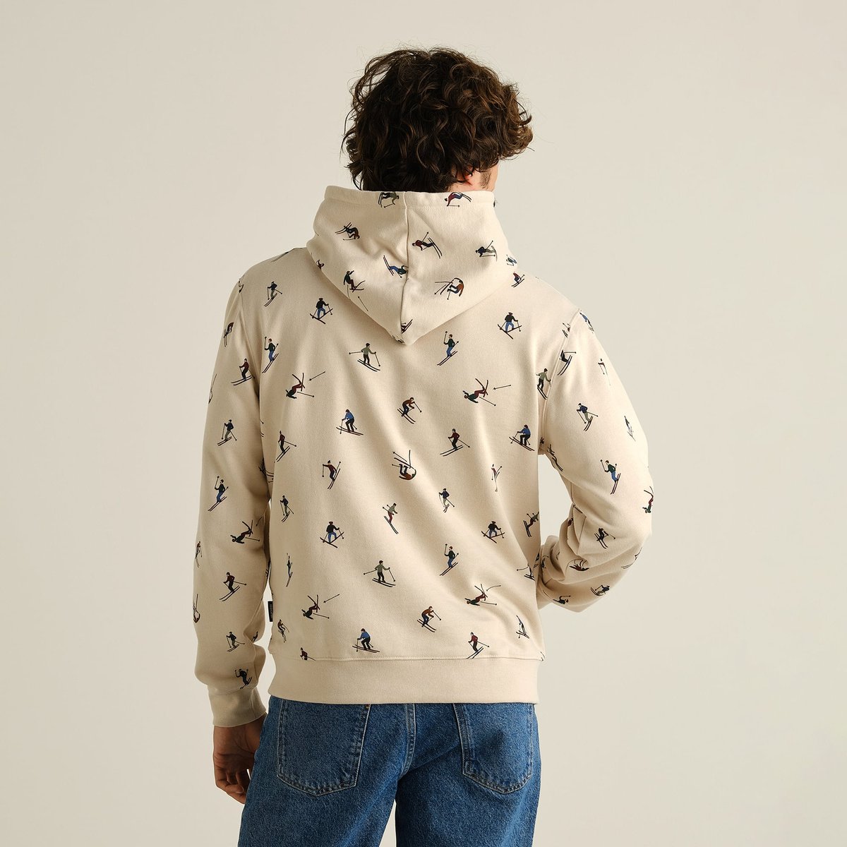 Back view of a model wearing the oat white Hoodie Falun Ski Team, featuring a playful all-over print of skiers.