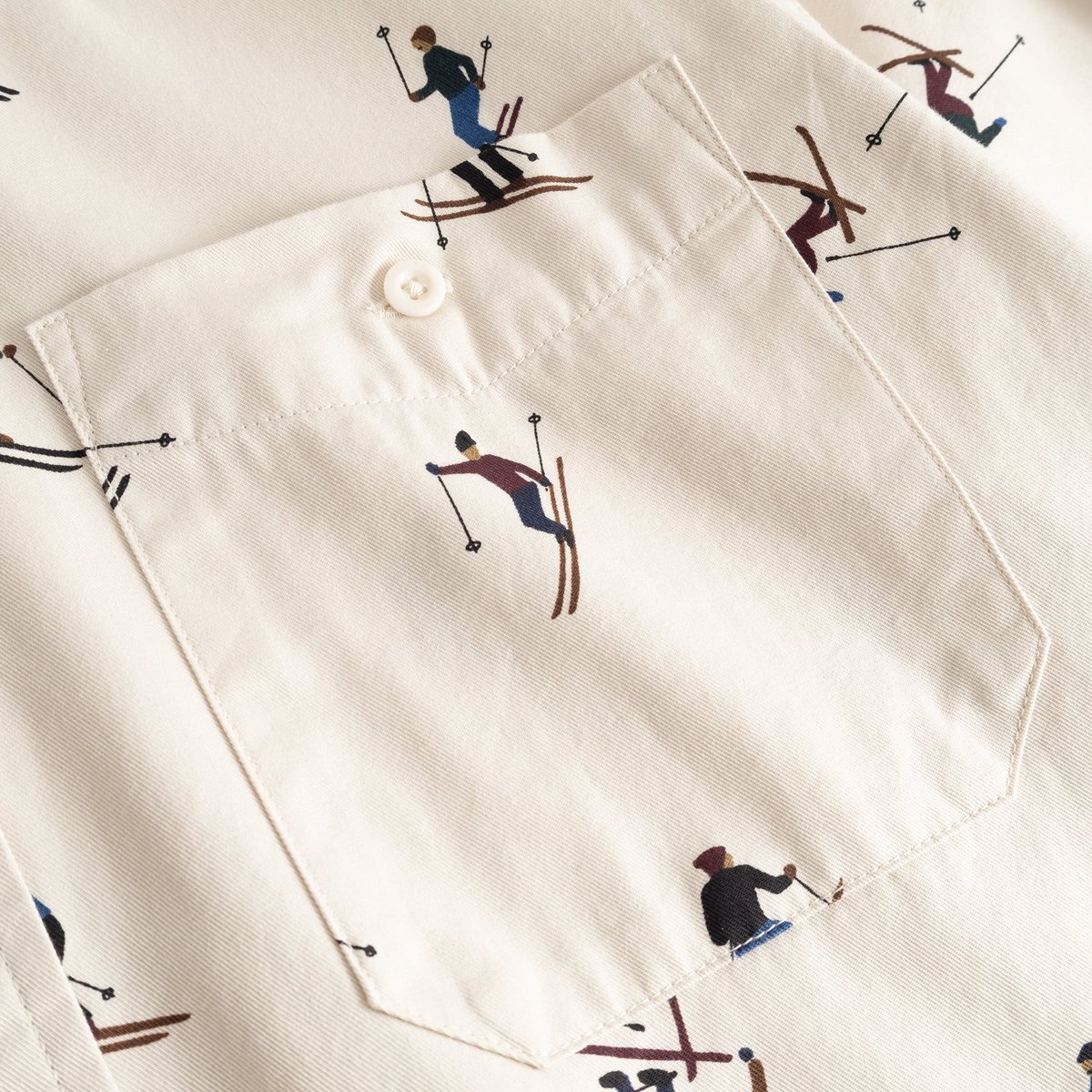 Close-up of the chest pocket on the Shirt Lunnevik Ski Team, showing the playful skier all-over print on an oat white fabric.