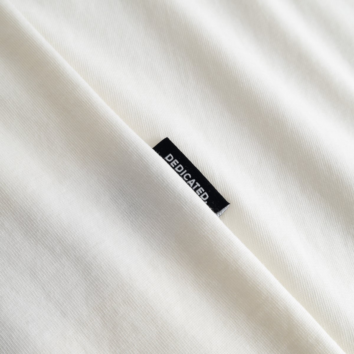 Close-up of the Dedicated brand label on the side of the soft, off-white T-shirt Stockholm Painted Side Bike.