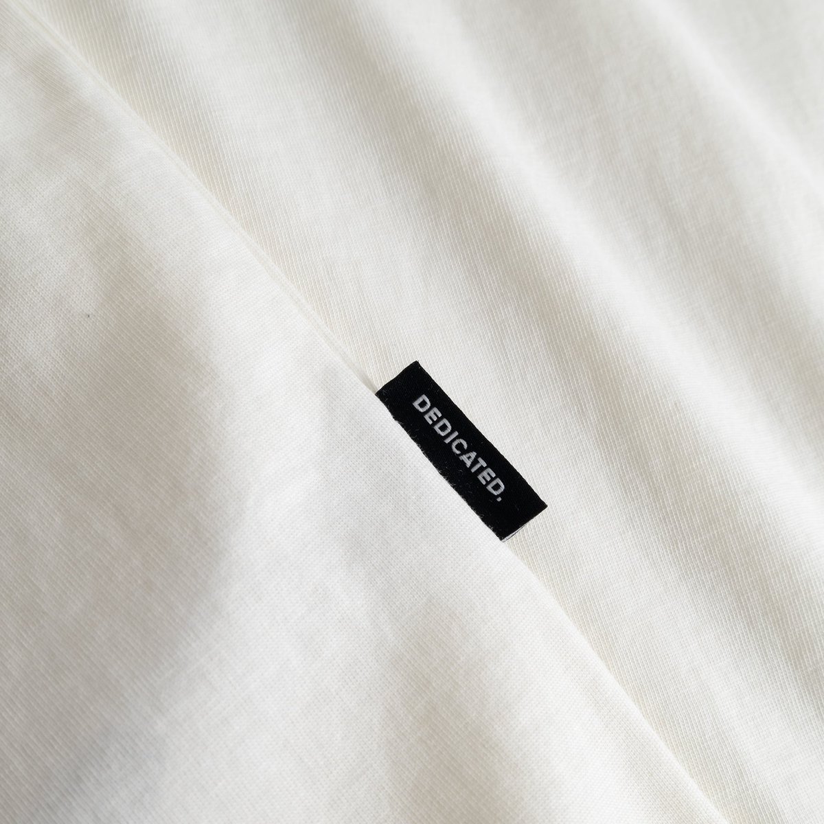 Close-up of the black Dedicated brand tab on the side seam of the off-white T-shirt Stockholm Camp Crew.