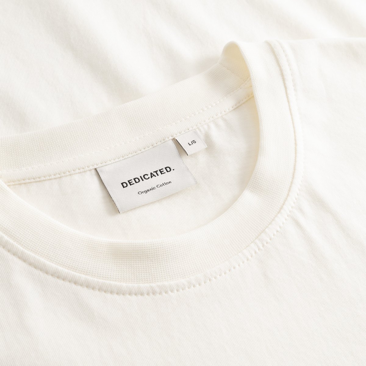 Close-up detail of the Dedicated brand label and ribbed collar on the T-shirt Stockholm Spilled Off-White.