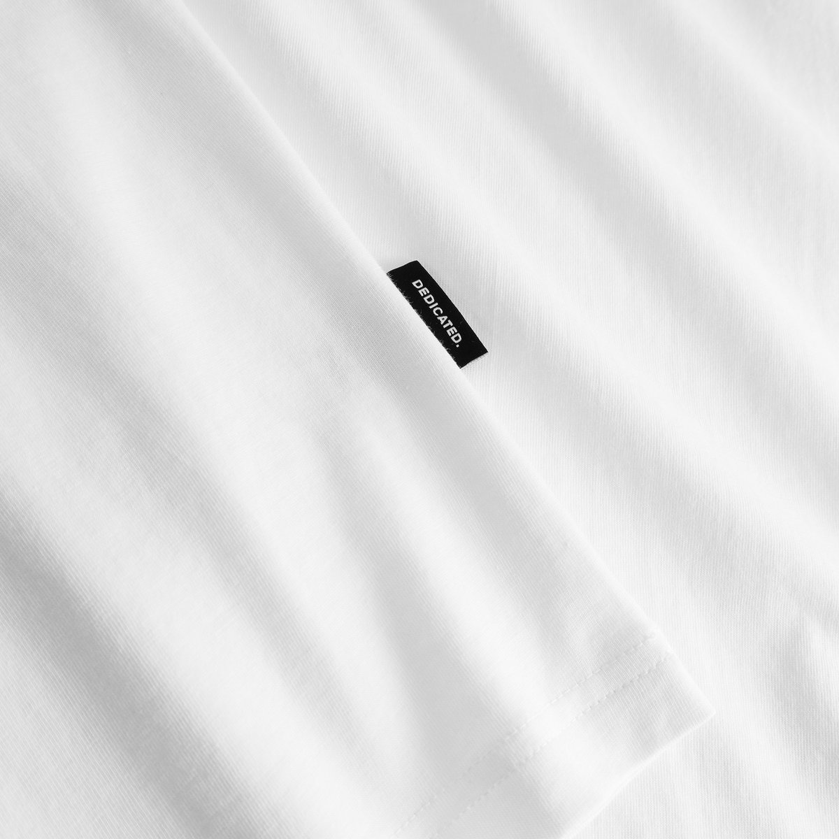 Close-up detail shot of the Dedicated brand tag on the sleeve of the white Stockholm T-shirt.