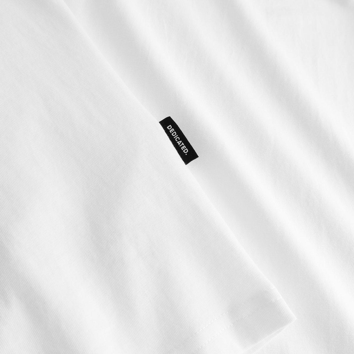 Close-up detail of the black Dedicated brand label on the sleeve of the white T-shirt Stockholm Race Bike EMB.
