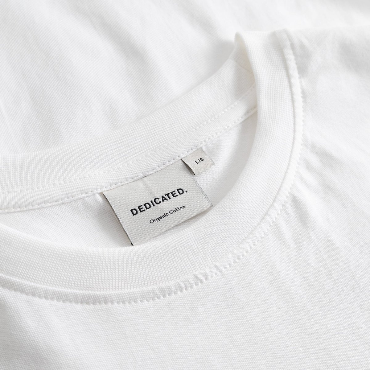 Close-up of the Dedicated brand label and ribbed collar on the white Stockholm Camp Crew t-shirt.