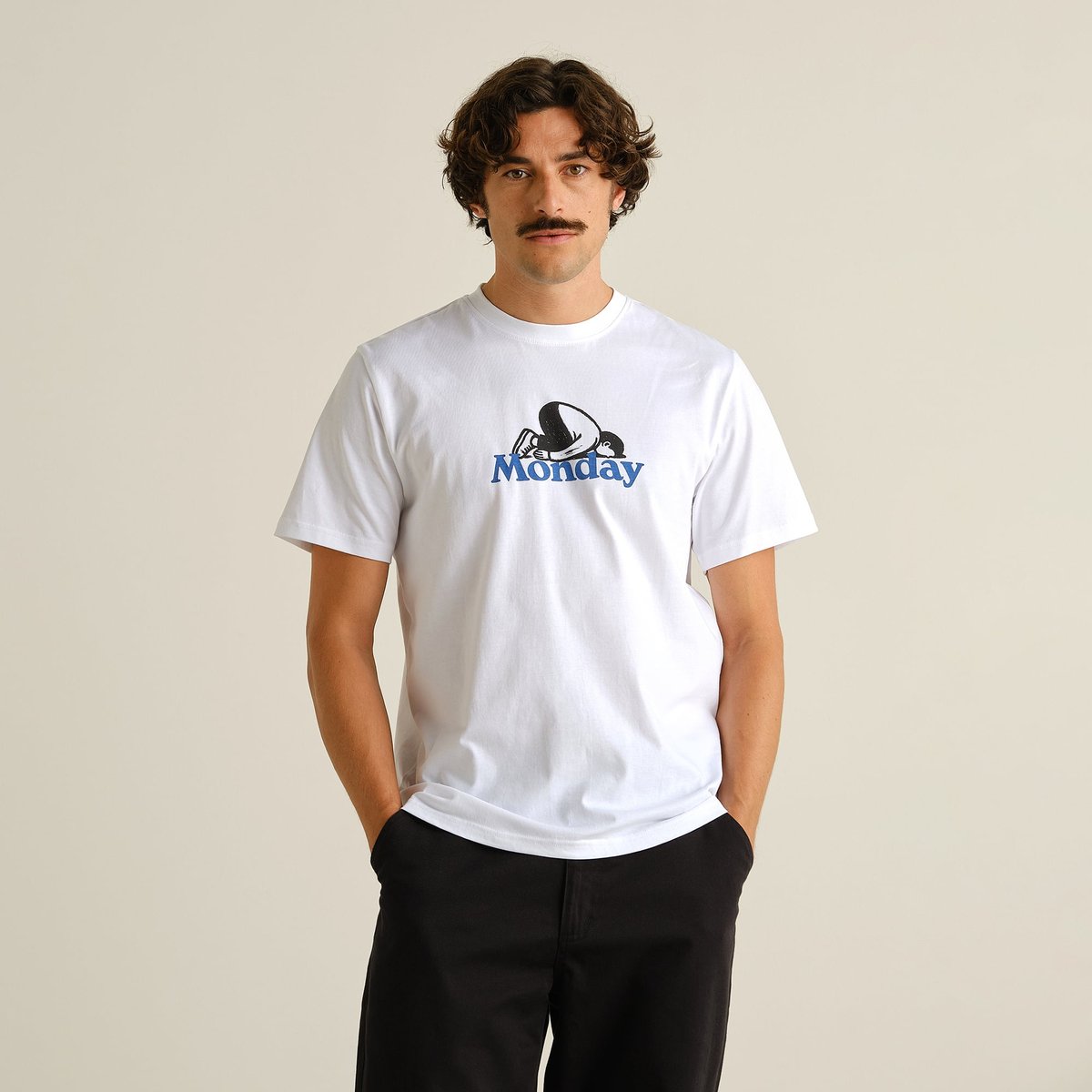 A male model with a mustache wears the white T-shirt Stockholm Monday Crash, featuring a tired character lying on 'Monday'.