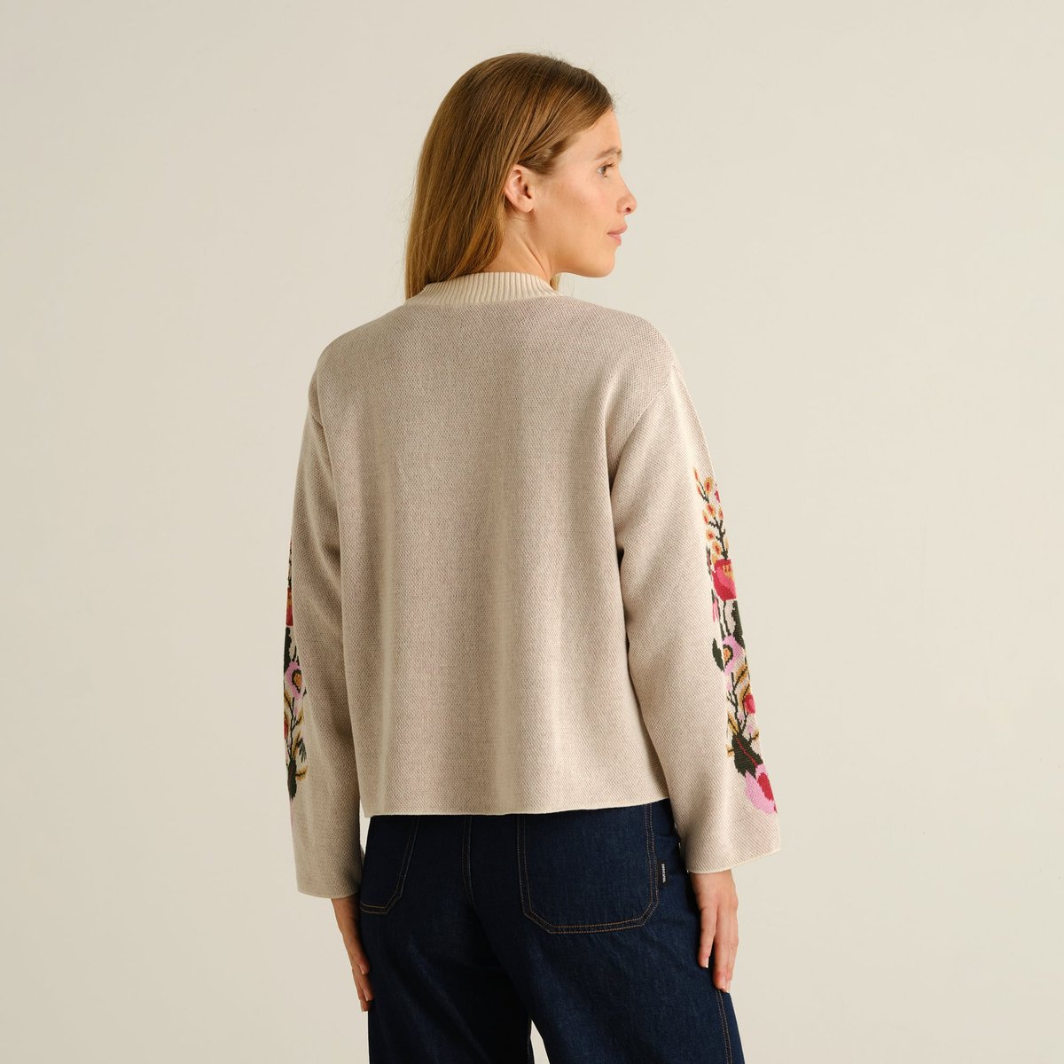 A woman models the back of the vanilla white Limhamn sweater, featuring a vibrant folk flower pattern on the sleeves.