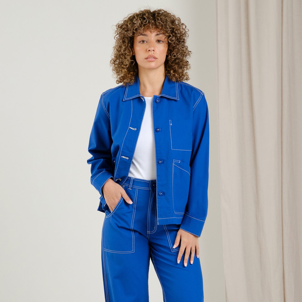 A model with curly hair wears the vibrant blue Stiby Canvas jacket and matching trousers with contrast stitching.