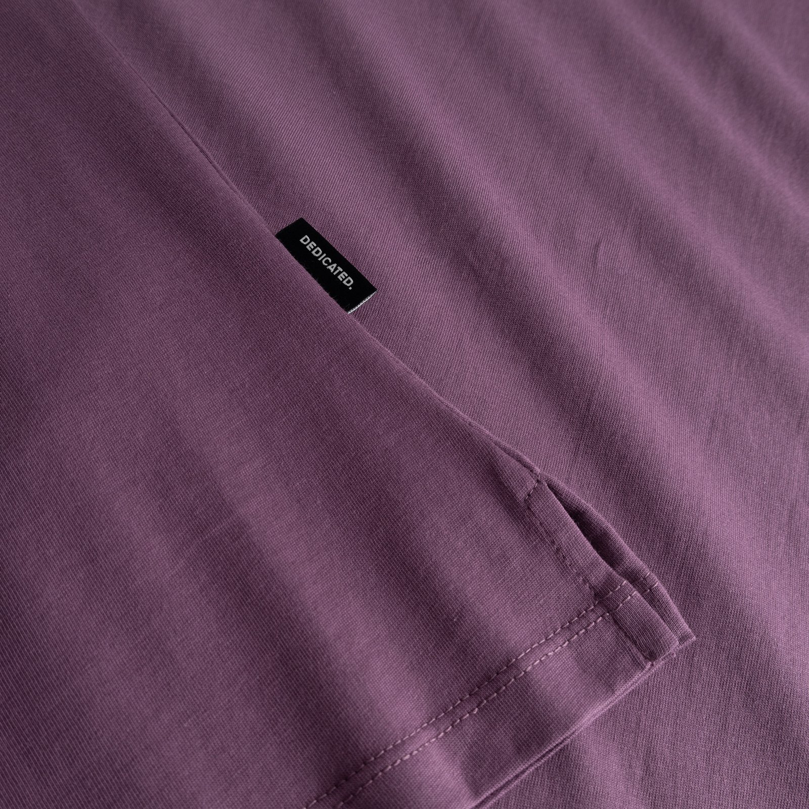 A close-up of the berry purple T-shirt Visby, showing the folded sleeve and the small black Dedicated brand label.