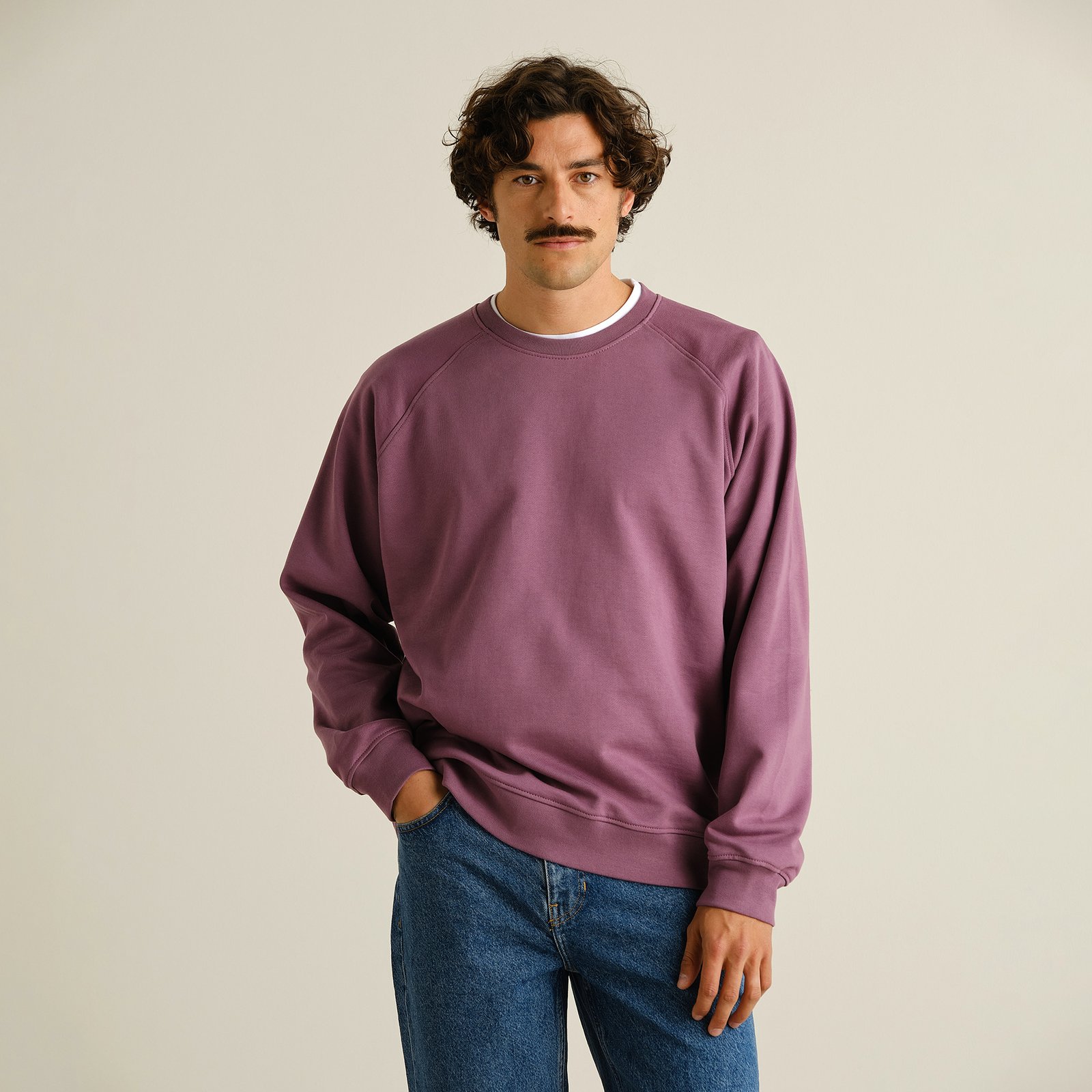 A man with curly hair and a mustache poses in a berry purple sweatshirt and classic blue jeans against a studio background.