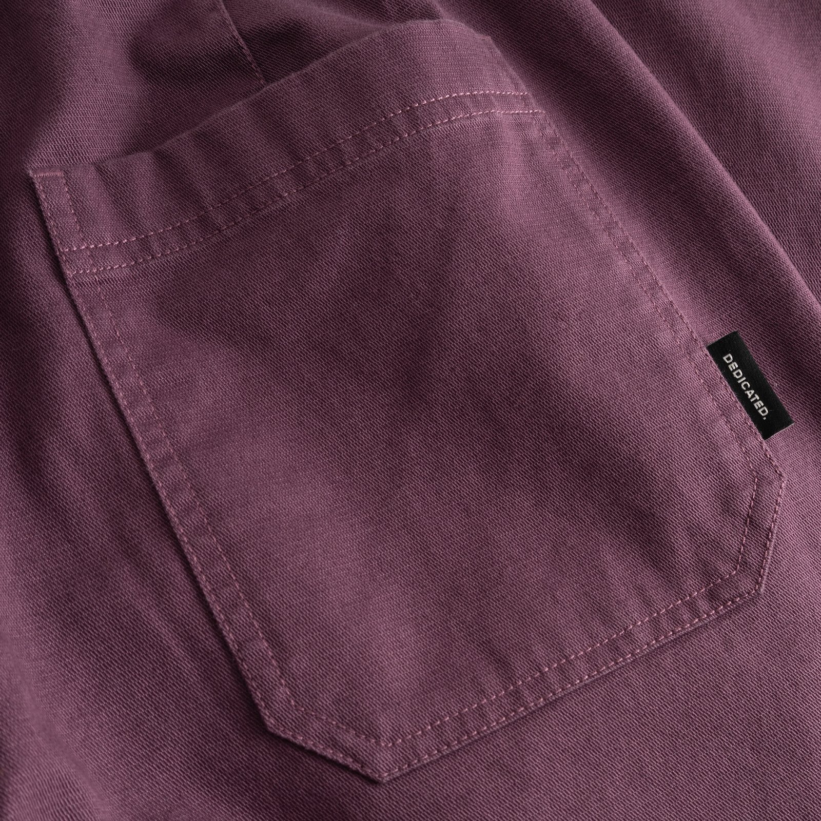 Närbild på bakfickan på Vara Workwear-byxorna i Berry Purple, med en svart Dedicated-etikett på sidan.