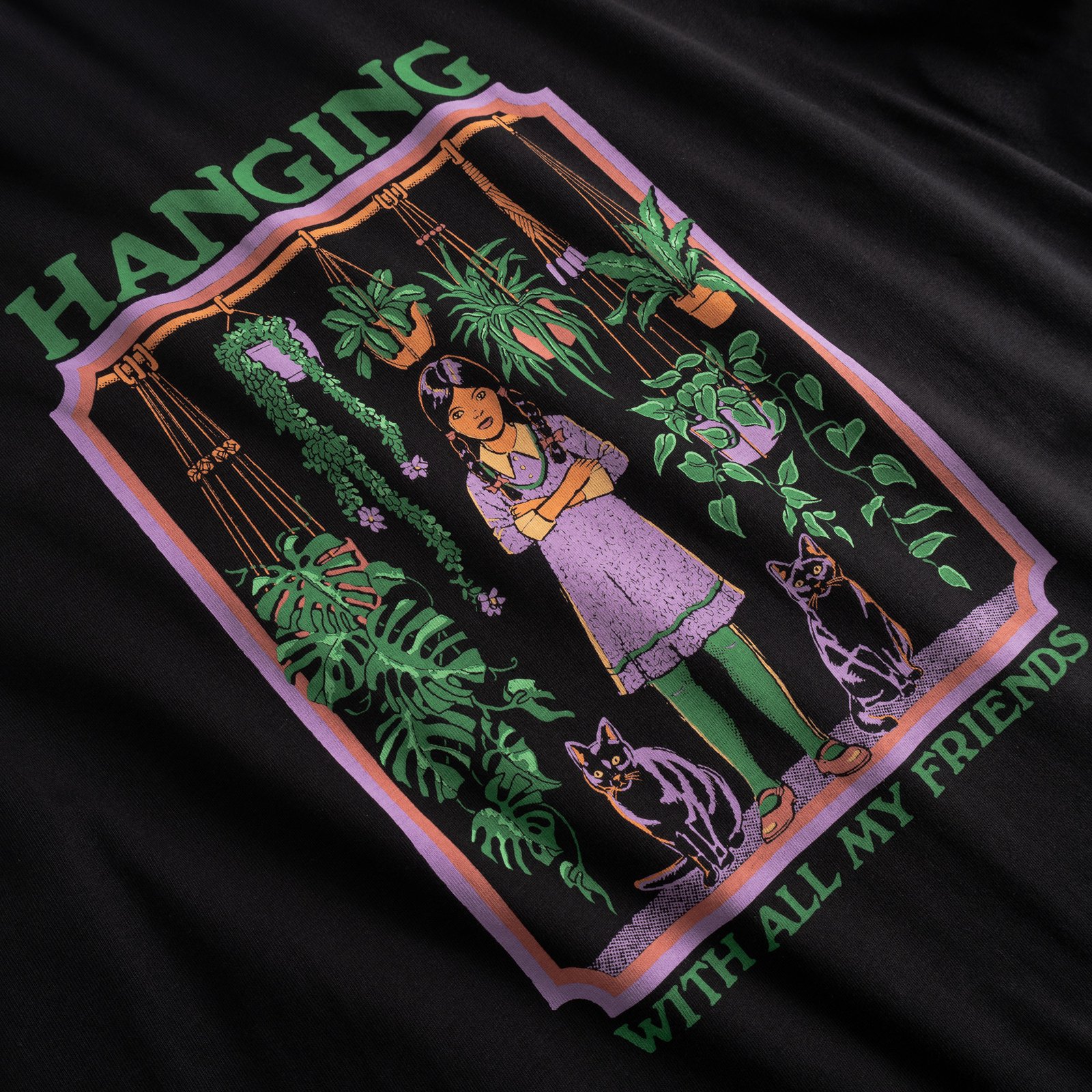 Close-up of the quirky 'Hanging With All My Friends' print, featuring a girl with her cats and plants, on a black t-shirt.