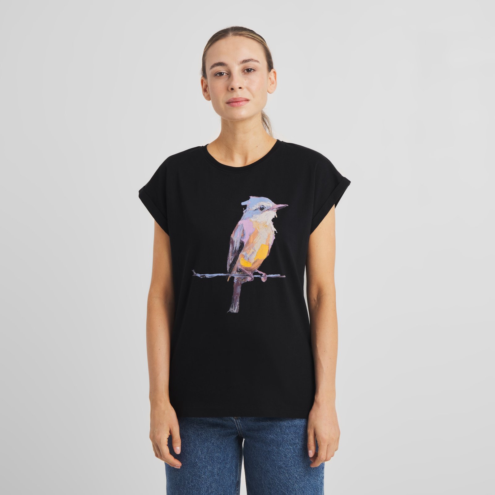Front view of a woman wearing the black T-shirt Visby Painted Bird, styled with blue jeans against a grey background.