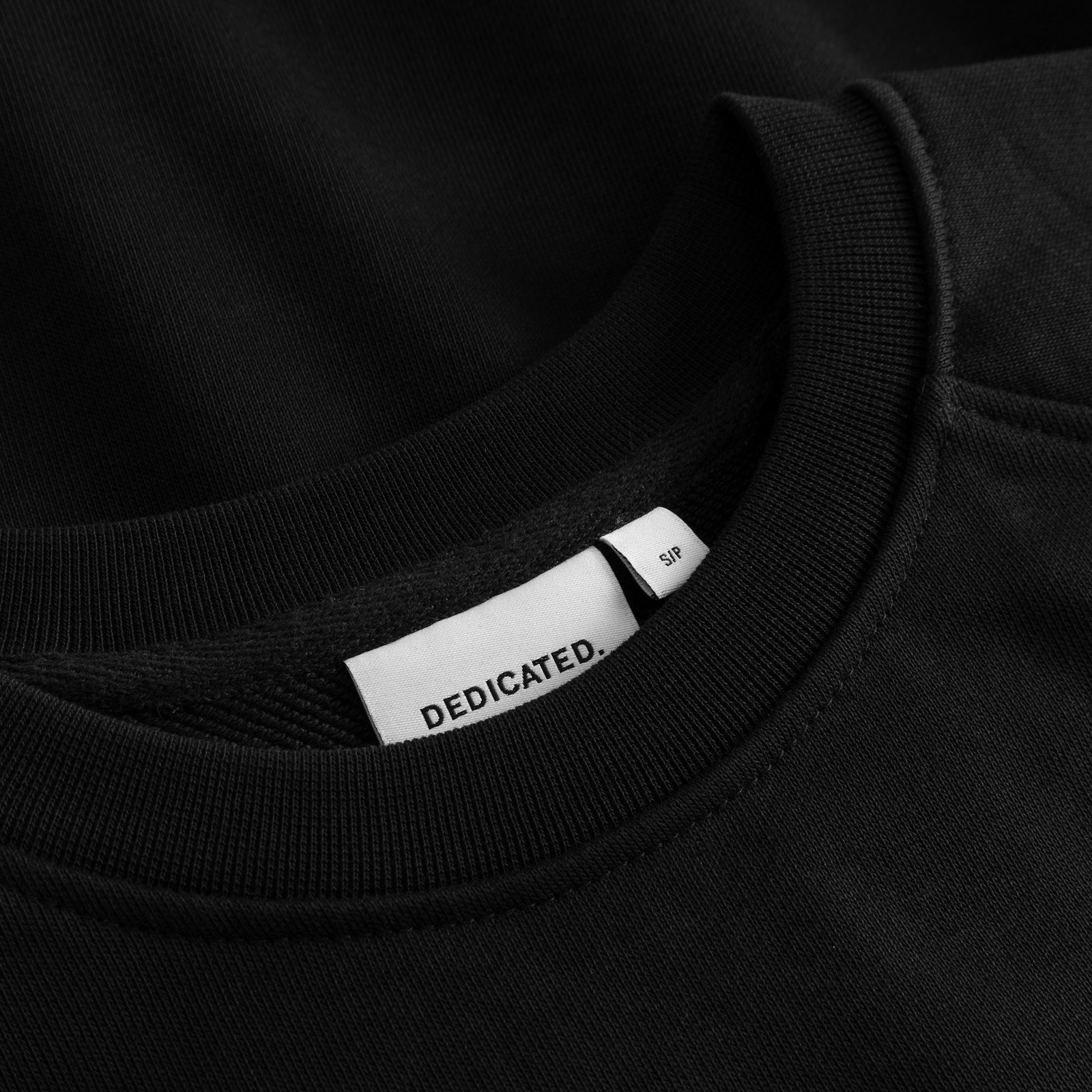 Close-up of the Dedicated brand label and ribbed crewneck on the black Sweatshirt Ystad Painted Swans.