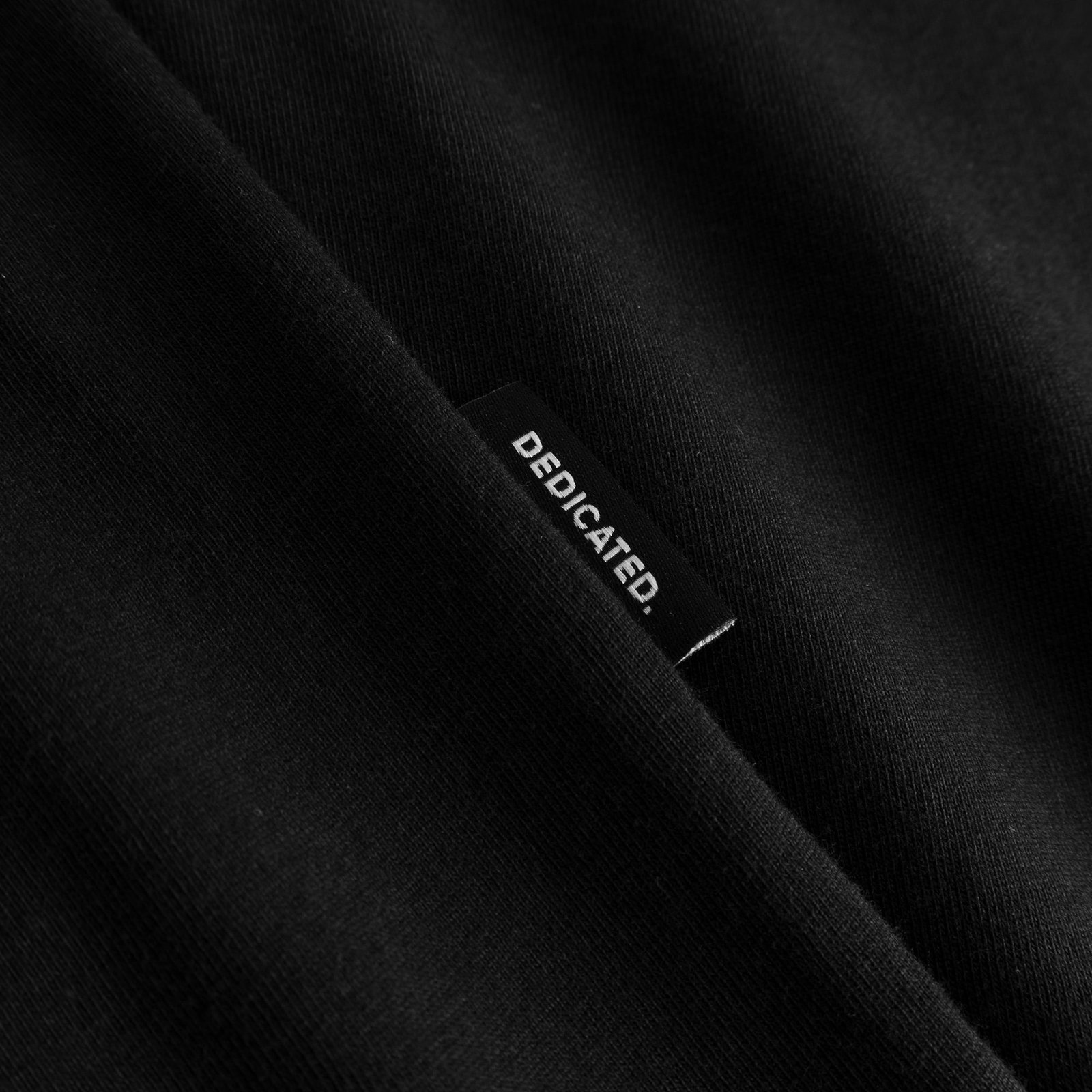 Close-up of the Dedicated brand label on the side seam of the black T-shirt Stockholm Apres Ski Instructor.