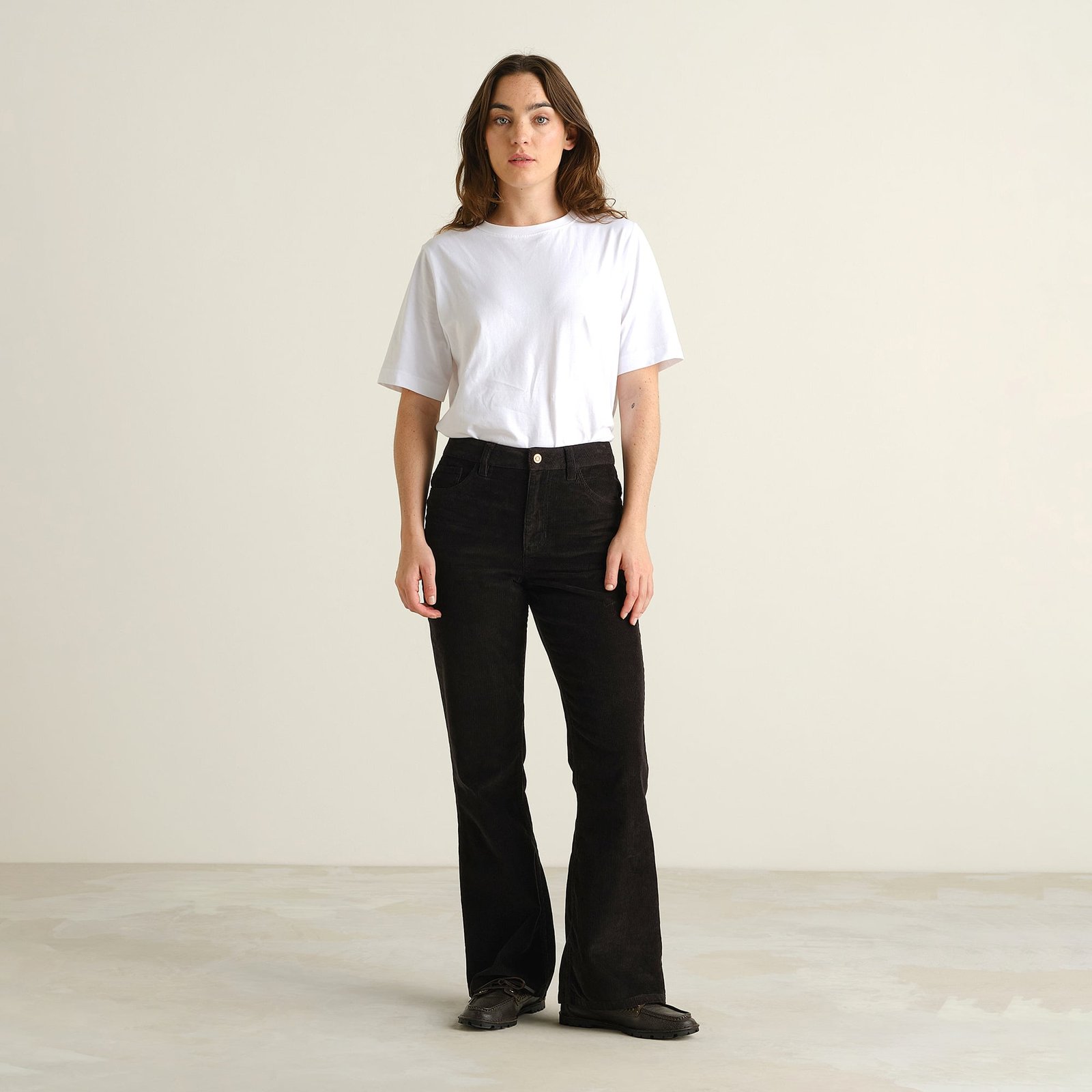Model in Frontalansicht trägt die schwarze Cordhose Ryda im Bootcut-Stil zu einem weißen T-Shirt.