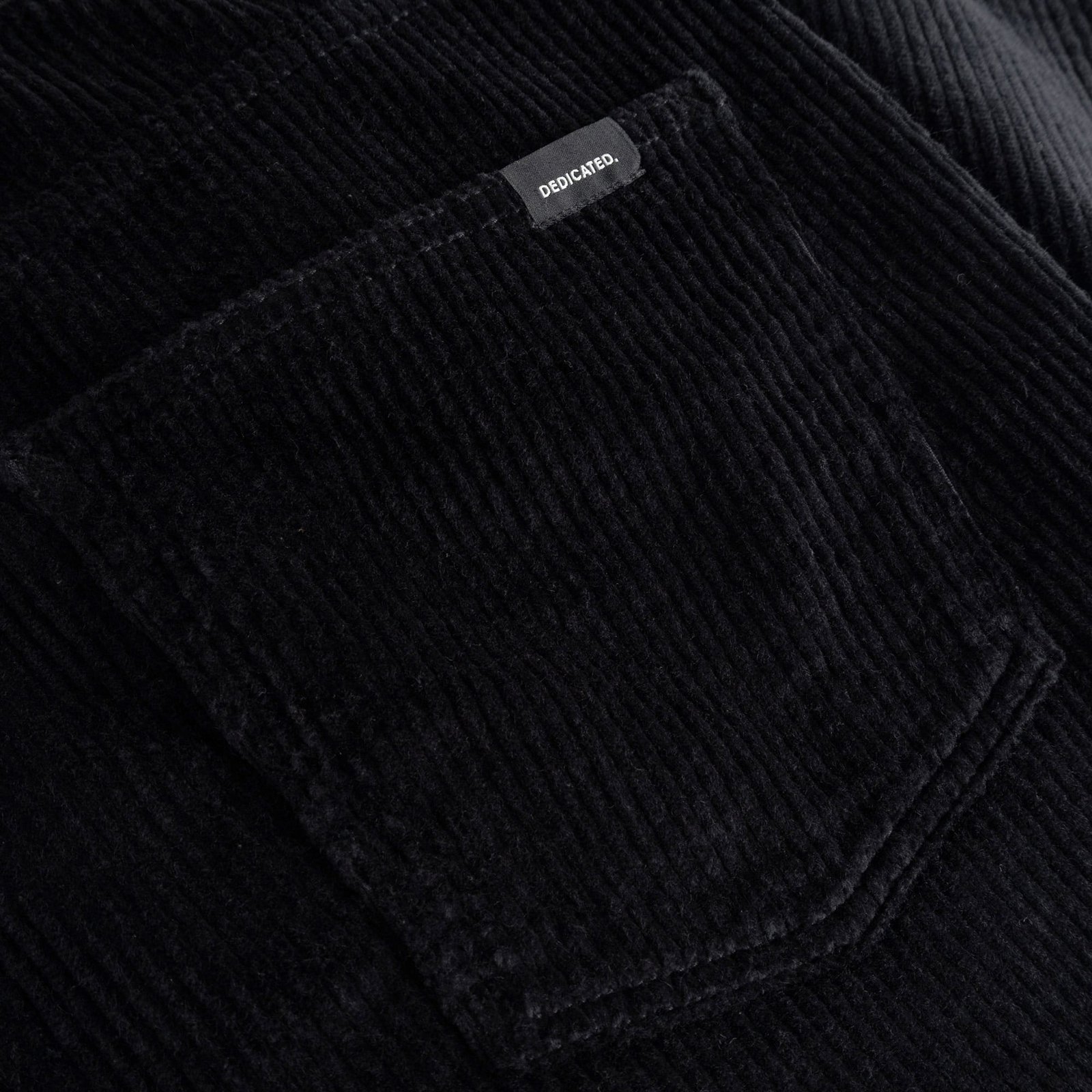 A close-up shot of the rich black corduroy fabric and back pocket of the Hamburg pants, showing the Dedicated brand label.