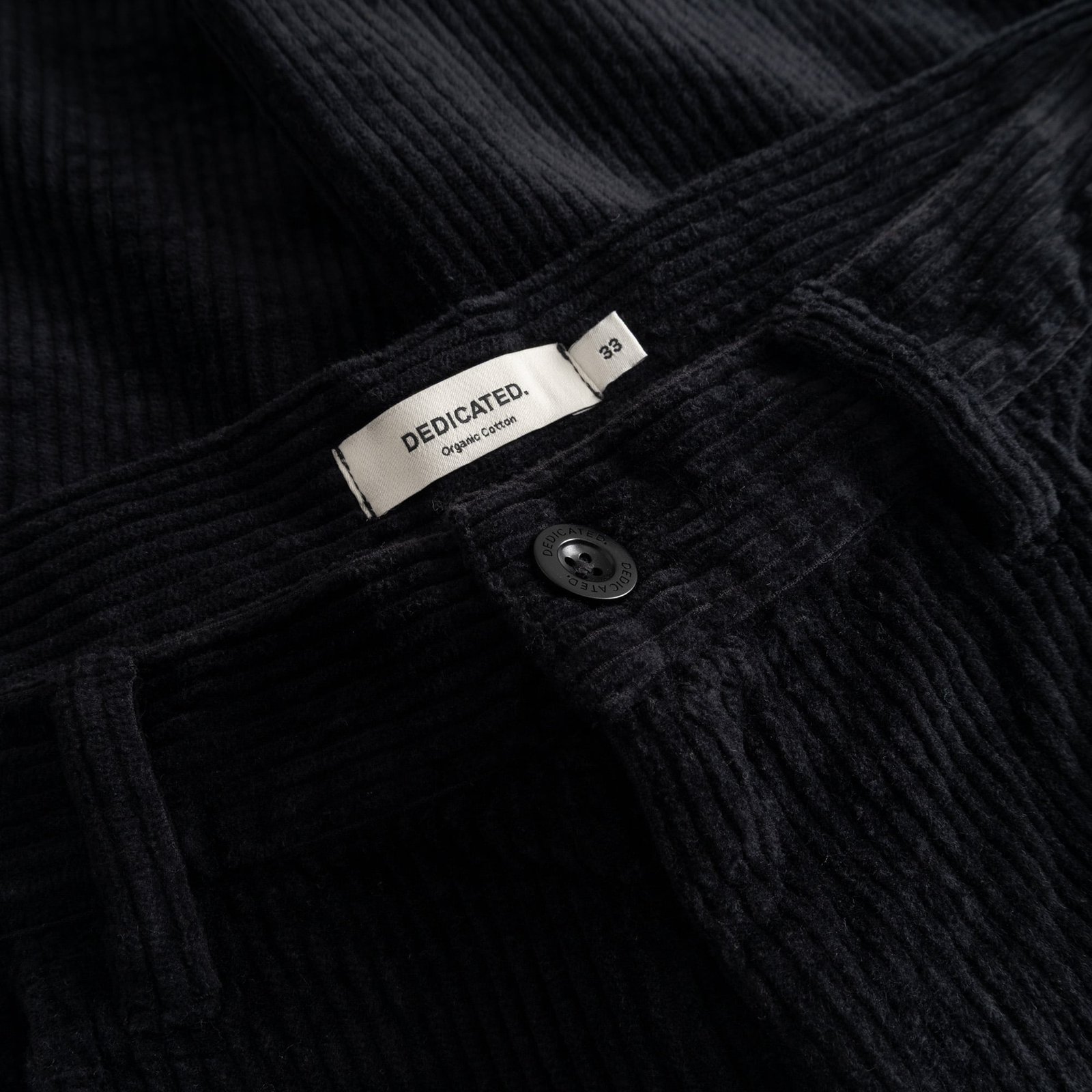 Close-up of the waistband on the black Hamburg Corduroy Pants, showing the Dedicated brand label and button detail.