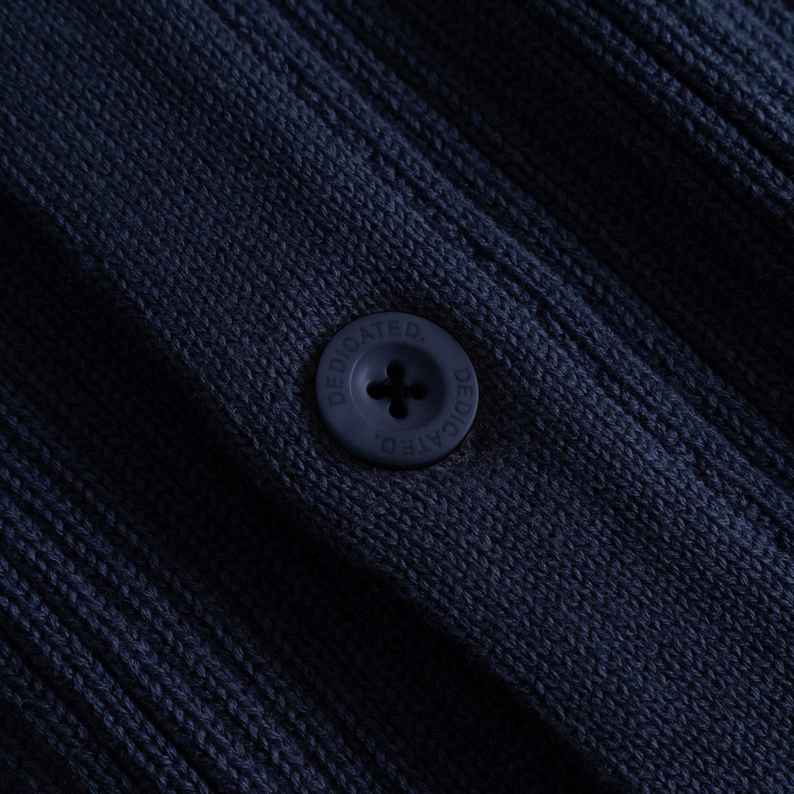 Närbild på den grovt ribbstickade texturen och en knapp med logotyp på den marinblå Cardigan Huuki.